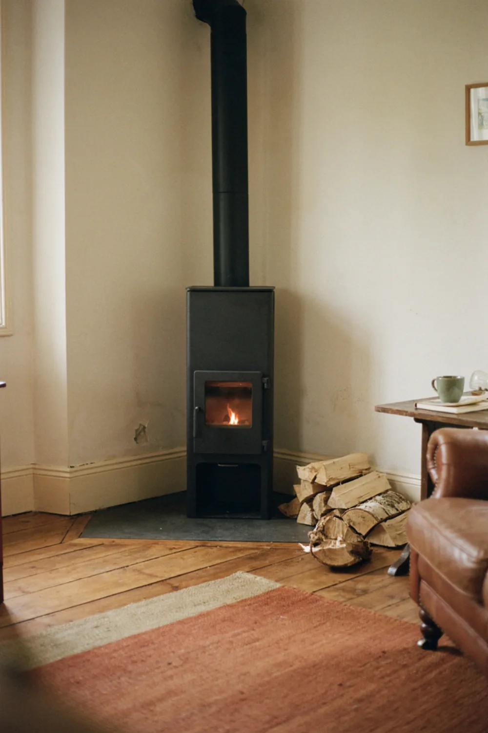 A slim black freestanding wood-burning stove style heater in the corner of a cozy small living room with a wooden floor